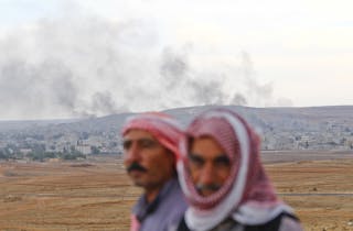 Turkish Kurds stand in front as somoke rises from the Syrian town of Kobani