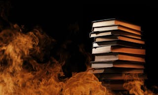 Conceptual image of a tall stack of hardcover books in a burning fire with flames and smoke swirling around them in a darkened r