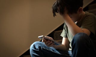 Cyber bullying concept. Young Asian preteen/teenage boy sitting at stair, covering his face with hand, other hand holding smartp