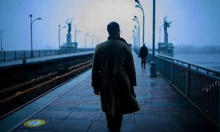 Back of cinematic young man in winter coat walking outside in urban city on bridge on a moody, foggy, winters night.