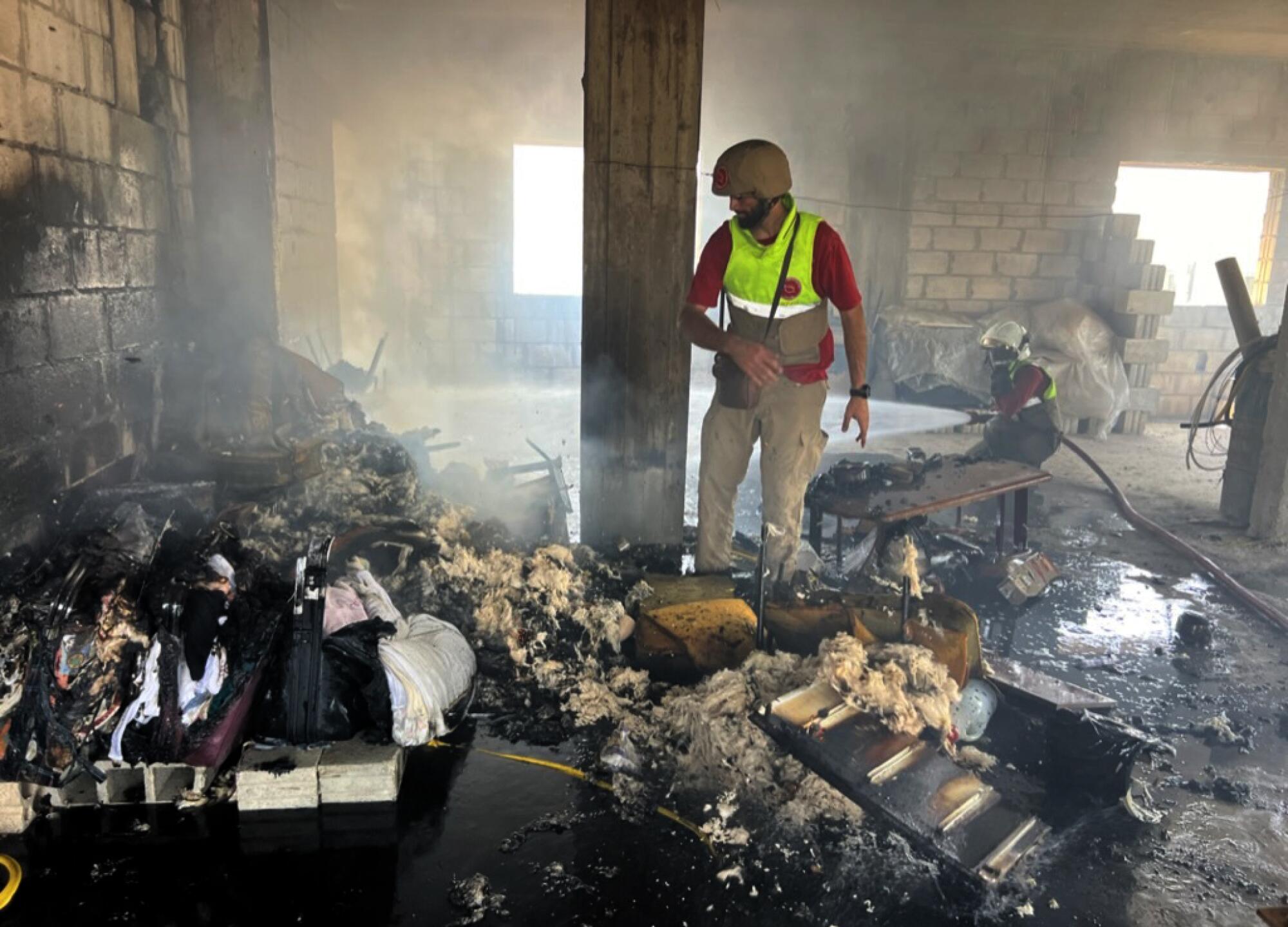 A man in a helmet and reflective vest stands amid smoking ashes inside a building.
