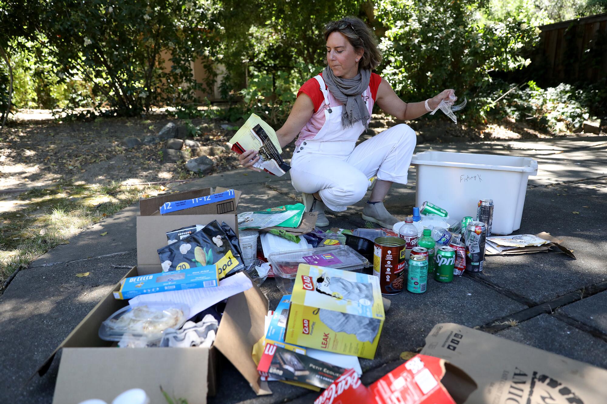 Susanne Rust wearing white overalls crouching down sorting groups of her family's trash on the ground outside.