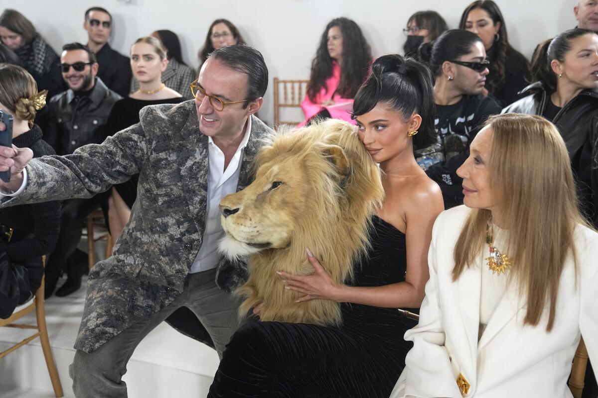 A man takes a selfie with a woman wearing a black dress featuring a lifelike lion's head while another woman looks on.