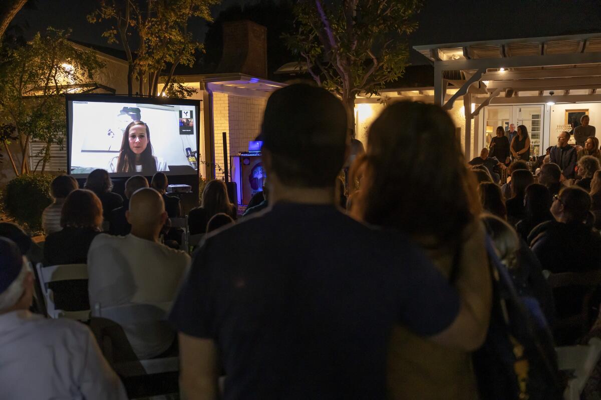 A large screen shows a woman while a crowd watches outdoors. 