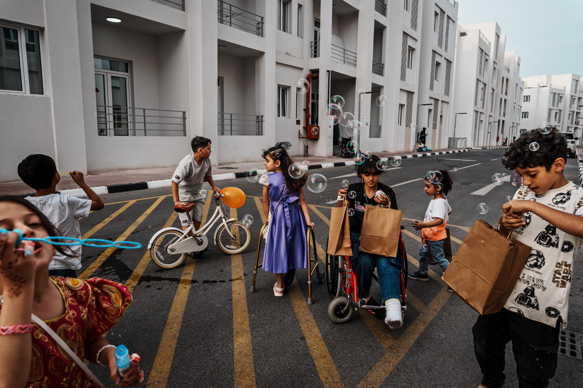 A group of children, including one in a wheelchair, holding bags, play on a street near a multistory building 
