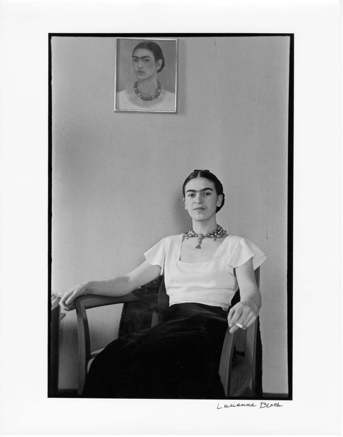 A youthful Frida Kahlo sits smoking in a bare room below one of her self-portraits in a vintage black-and-white photo.