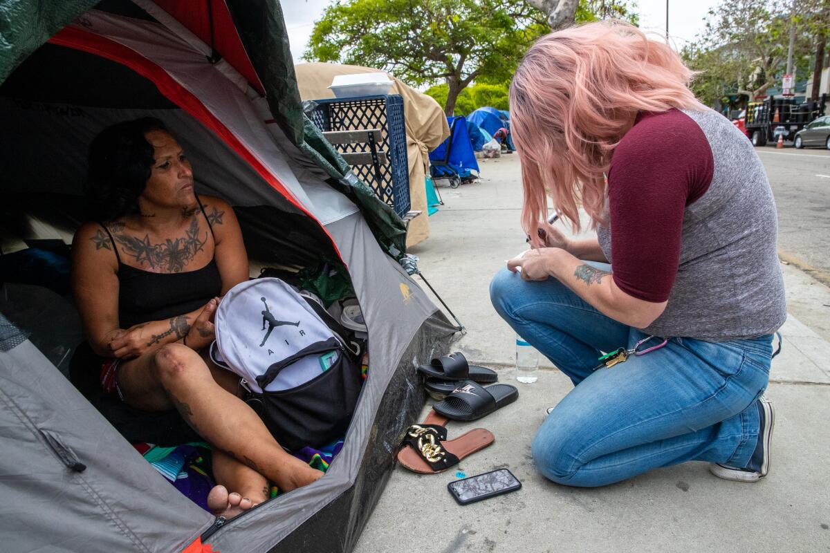 A woman sits in a tent on a city sidewalk while another woman kneels outside the tent taking notes