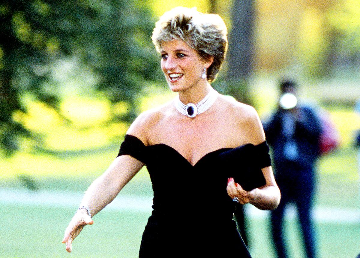 Princess Diana walking in a black dress and pearl choker holding a black purse on her left with the other arm thrust forward