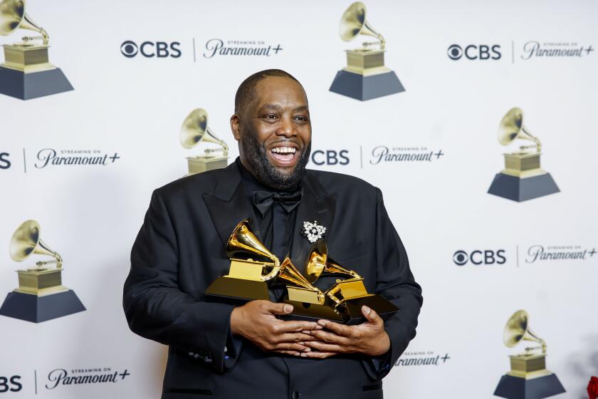 Killer Mike in a black suit holding three golden Grammy Awards in his arms as he smiles and poses for pictures