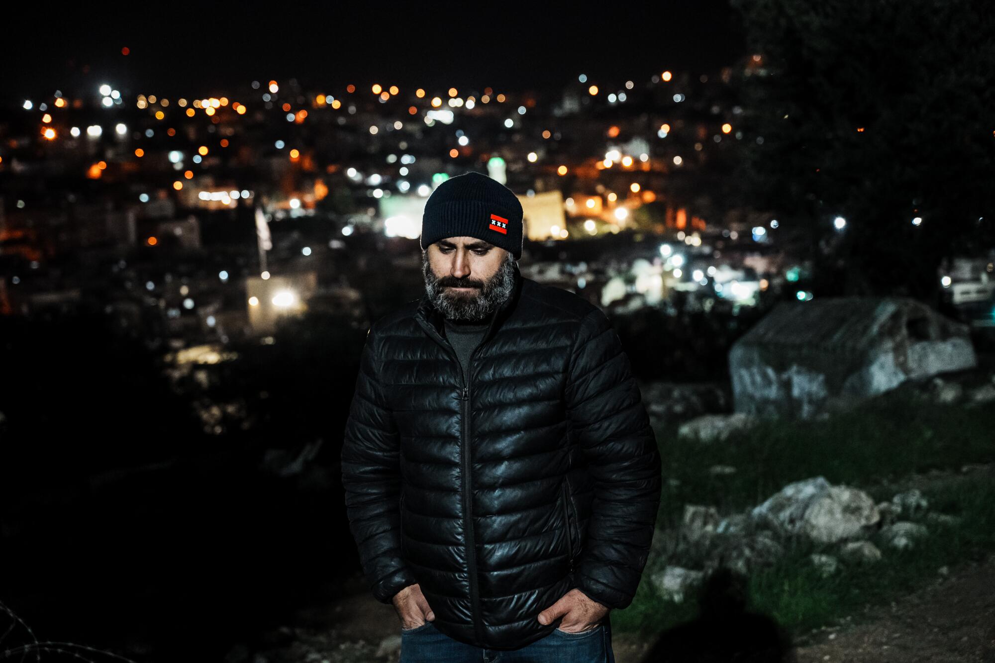 A bearded man in a hat and puffer jacket standing, hands in his pant pockets, on a hill in the dark, city lights behind him