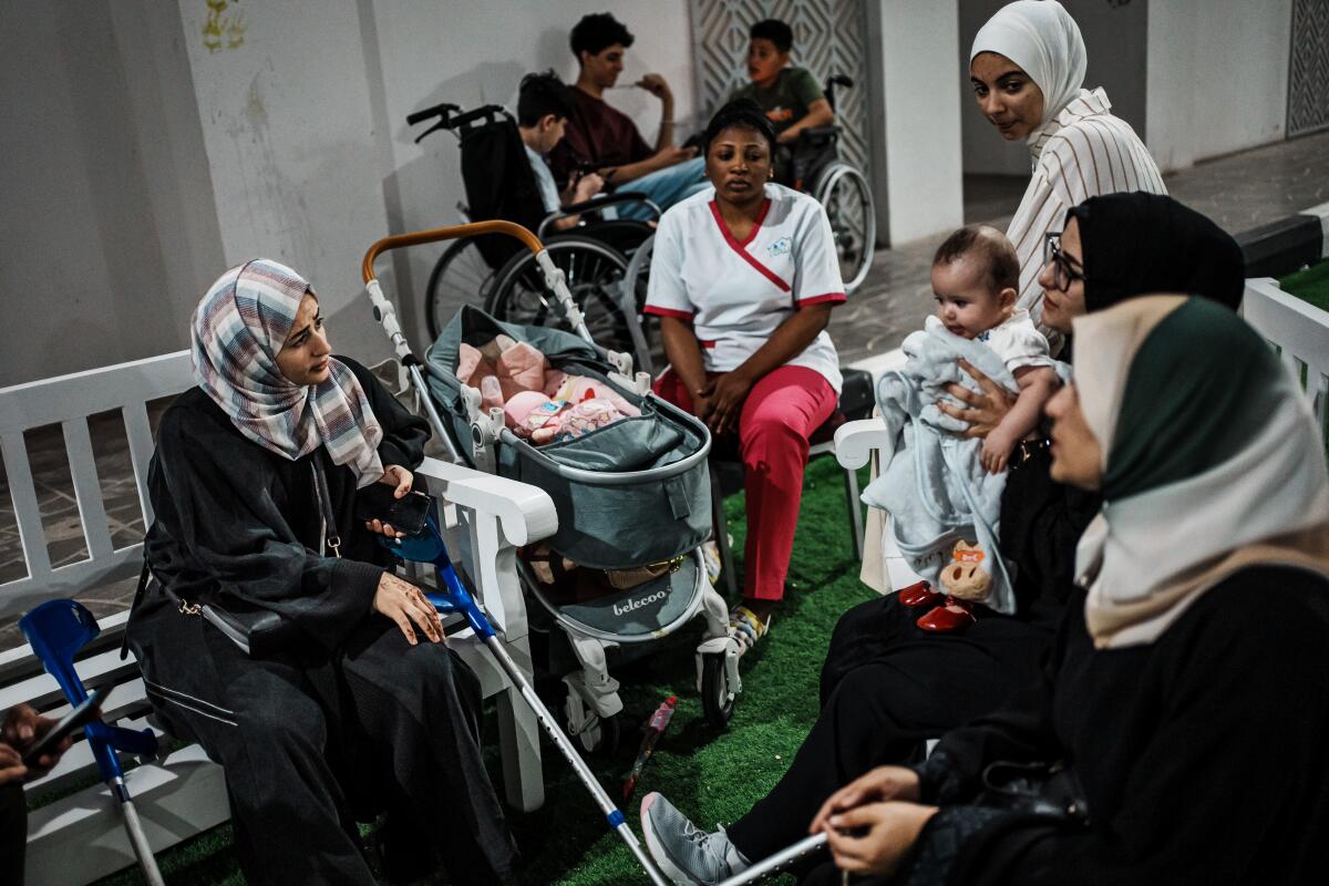 Women sit on benches and hold babies or watch children in strollers as people in wheelchairs sit and talk in the background.
