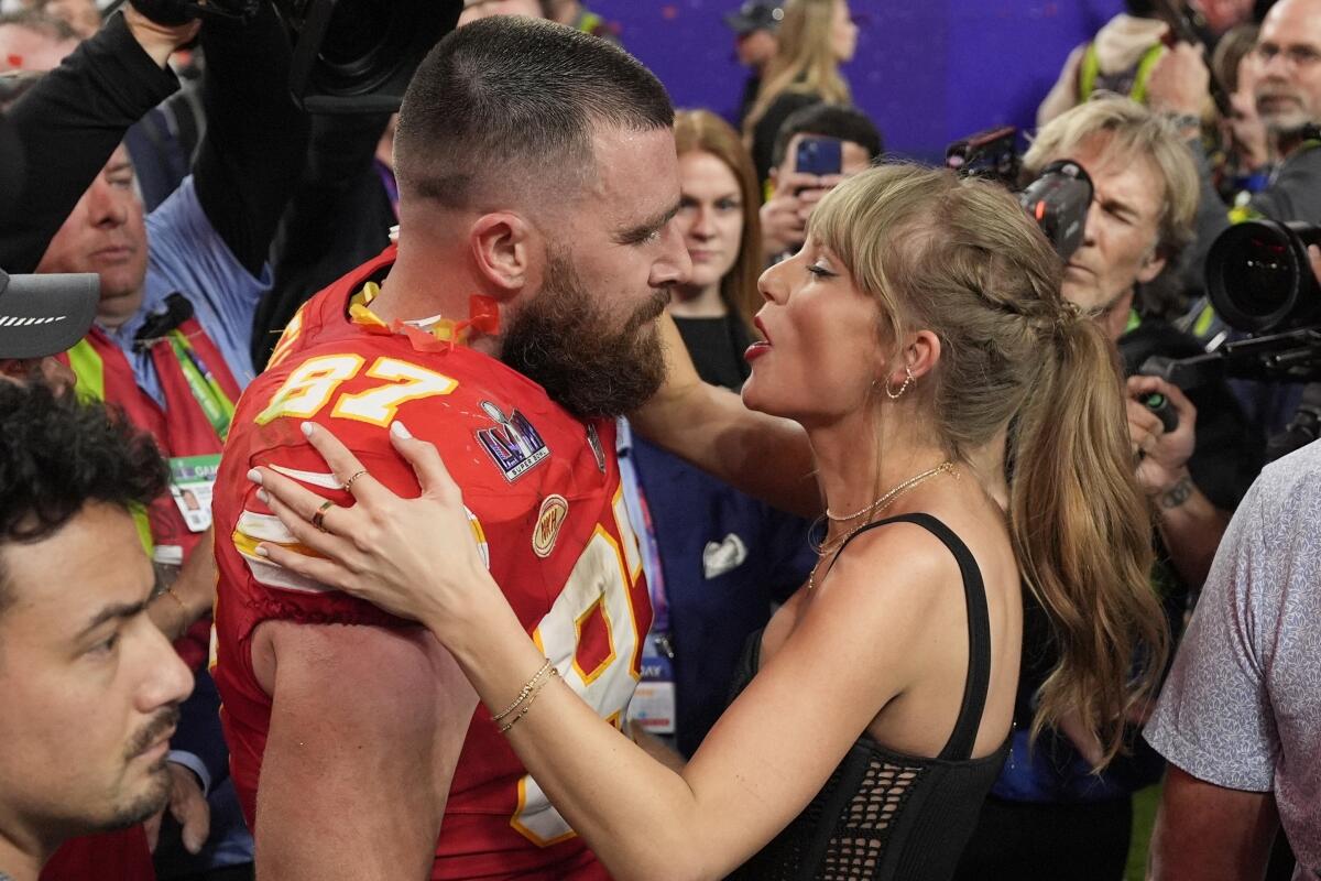 Taylor Swift embraces Travis Kelce wearing his Chiefs uniform on the field in front of men with cameras