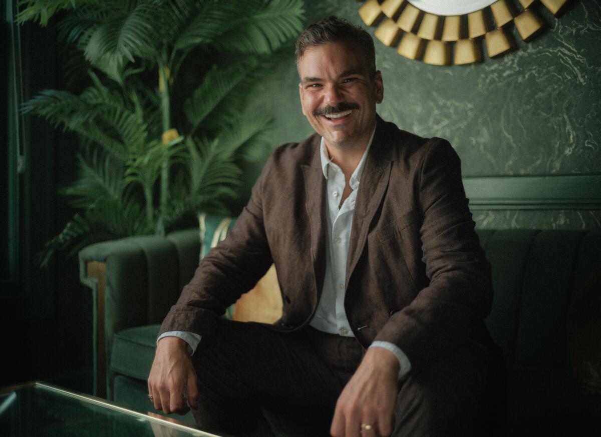 Ian Karmel, with a mustache and brown suit, laughs while seated on a green couch.