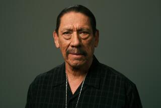 Danny Trejo wearing a black v-neck shirt and a thin necklace looking forward against a grey-ish backdrop