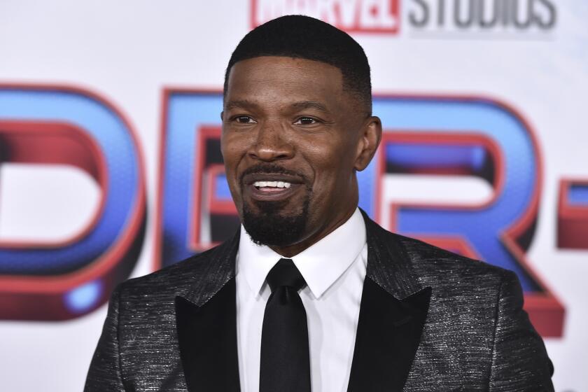 Jamie Foxx in a dark patterned suit and tie smiling for a  a Spider-Man red carpet