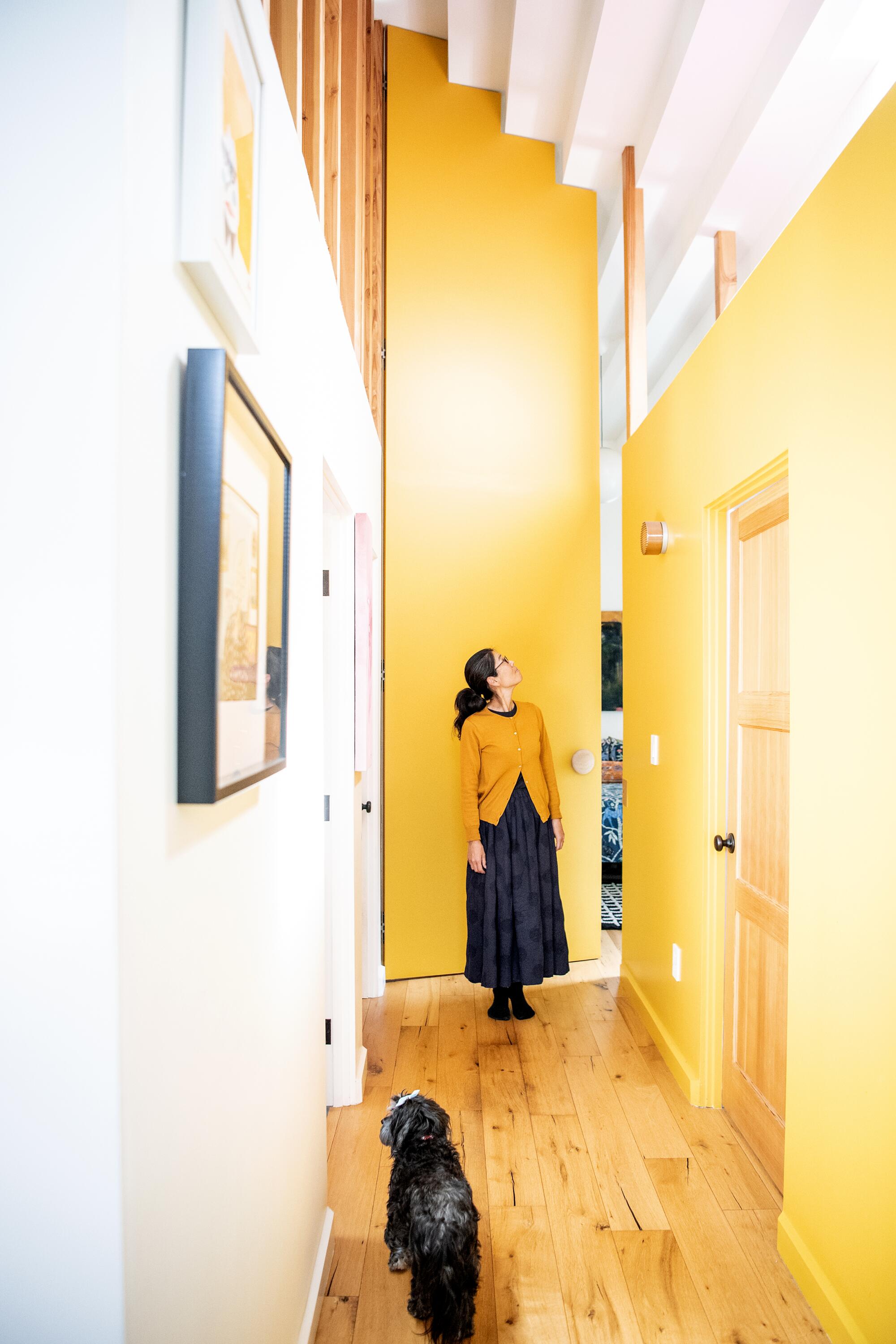 A woman stands in a hallway, in front of a tall door that's ajar, with a small black dog.