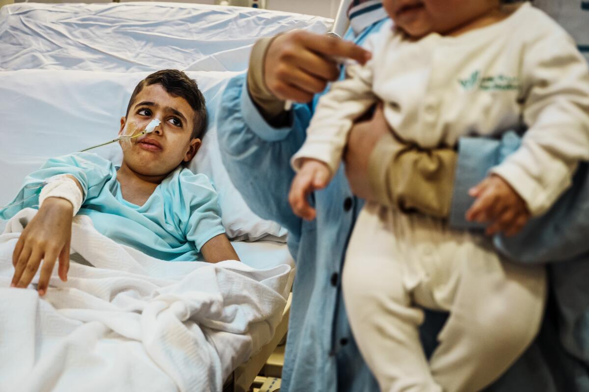 A 10-year-old Palestinian boy lies in a hospital bed, while in the foreground his mother holds a baby. 
