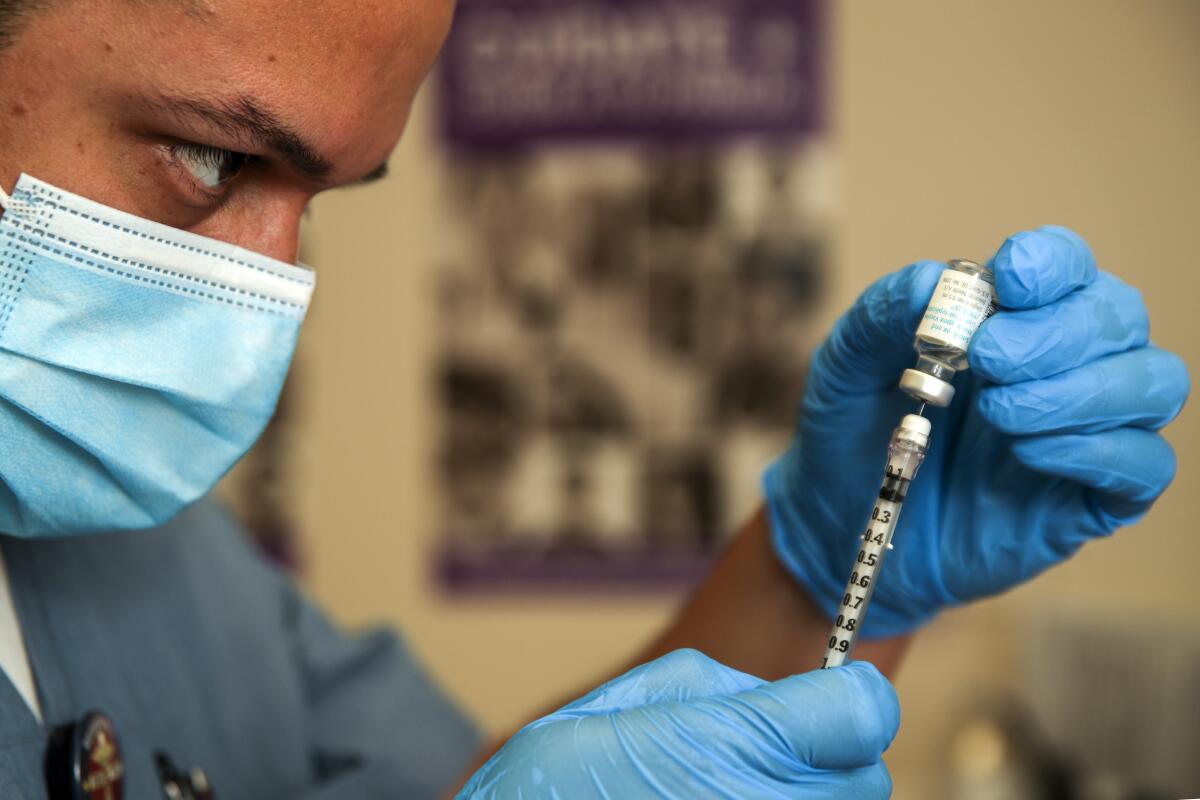 Close-up of a person in mask and other medical gear, holding a vial and filling a syringe.