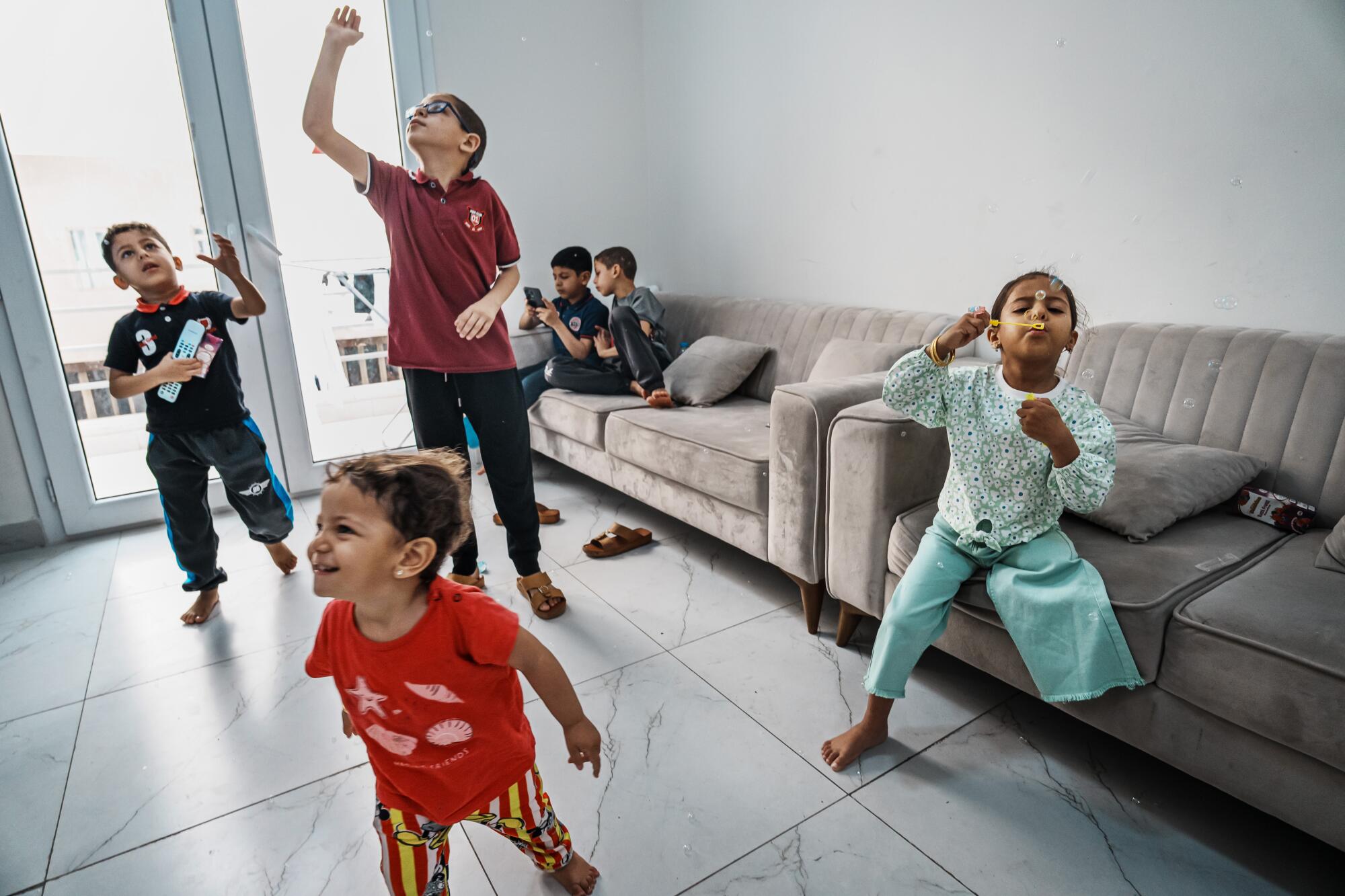 Six children blow bubbles while playing on a tile apartment floor or on couches. 