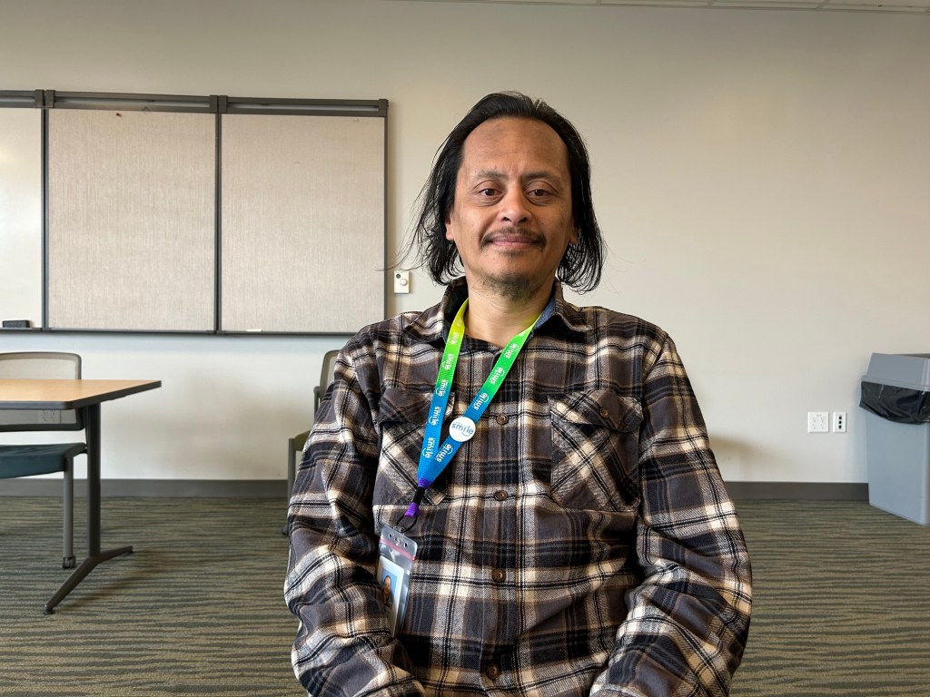 Derrick Cordero sits in a chair in the center of the photograph. He rests his hands on his knees and looks directly towards the camera with a smile.