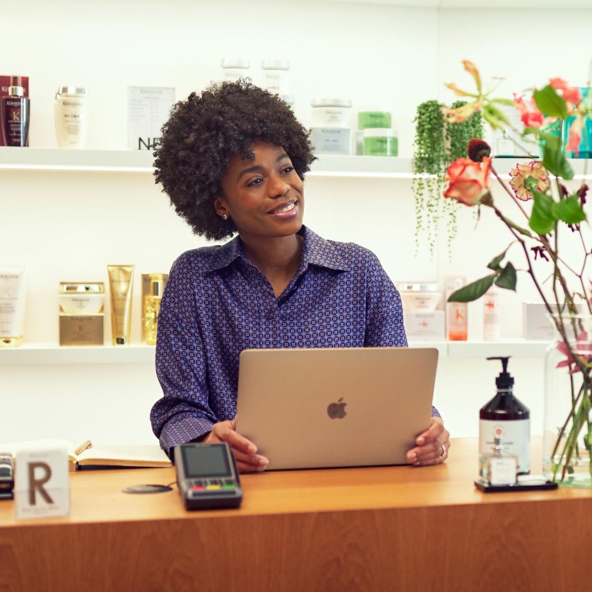 Smiling person using a laptop at a retail checkout counter with beauty products and a card payment terminal.