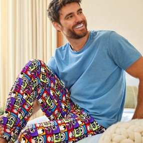 A happy man sitting on a bed wearing a blue t-shirt and Marvel pajama pants with Ironman's helmets and other avenger symbol details.