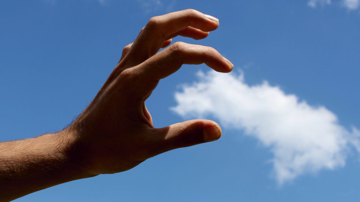 A hand acting as if it can grasp a cloud in the sky, shot from an angle to make it look real.