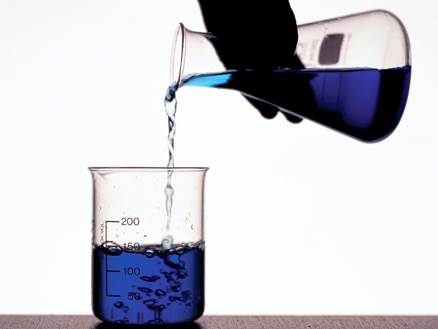 A person's hand pouring blue fluid from a flask into a beaker. Chemistry, scientific experiments, science experiments, science demonstrations, scientific demonstrations.