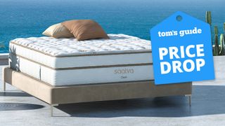 A Saatva Classic Mattress on a bed frame outdoors on a platform by the sea, with a blue price drop sale badge overlaid on the image