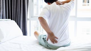 A man sits on the edge of his bed, with a hand held to his hip and neck.