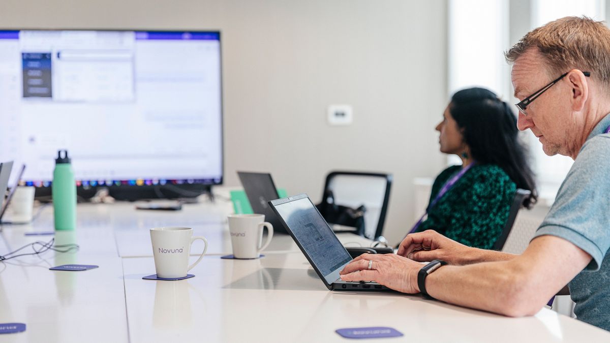 Novuna employees sat at a table in a boardroom. A man near the frame works on a laptop, while a woman sat behind him watches a large screen.