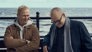 Johnny och Jan sitter bredvid varandra på en bänk vid havet och ser ut att vara på gott humör.