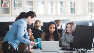 A female teacher explaining something to a small group of female students, who are sat around a desk looking at a laptop. All are sat in their school's computer lab.