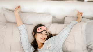 A woman wearing a pink sleep eye mask and silk pyjamas stretching her arms in bed waking up from quality sleep.