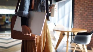 Return to office concept image showing young woman holding laptop and flask walking through an open plan office space.