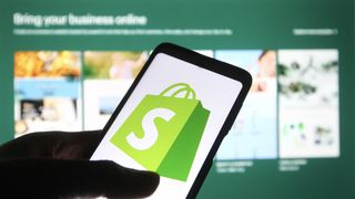 The Shopify logo on a smartphone in front of the background of the Shopify website