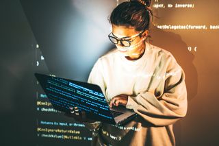 A woman stood typing on a laptop while in front of a projector casting an image of computer code