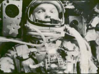 John Glenn, astronaut, is seen from the chest up, sitting in his spacesuit. Two tubes come from out of frame to connect to his helmet. His eyes look left, as the machinery on both sides of his head fills the rest of the photo.