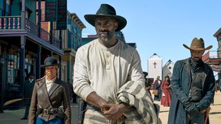 Idris Elba as Rufus Buck in The Harder They Fall as two men stand behind him in a Western town.