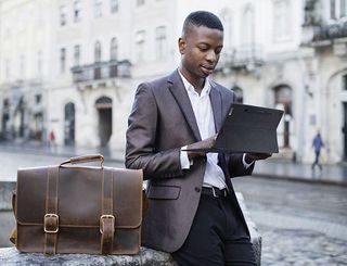 Ein Mann im Anzug hält das Lenovo IdeaPad Duet 5 in der Hand, während er an einem Brunnen lehnt.