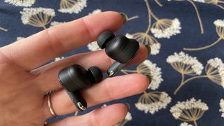 JBL Tour Pro 3 earbuds held in a hand, with navy floral background