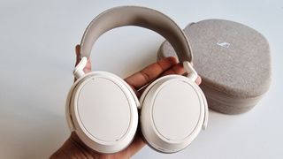 Bästa over-ear-hörlurar: En hand håller upp ett par beige Sennheiser Momentum 4 över ett vitt bord där hörlurarnas tillhörande fodral ligger.