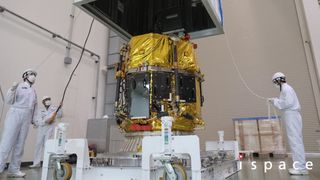 Three technicians in white coveralls and helmets hold lines connected to a large box hovering above a gold-foiled cylinder secured in the center.