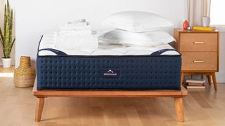 The DreamCloud Hybrid mattress shown on a light wooden bed frame and covered with white bedding in a white bedroom