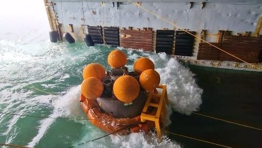 a large metal capsule floats in rough water using a giant orange floaty ring and six large orange floats near the top.