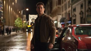 Tom Cruise as Jack Reacher looking at something off camera with a red car behind him. 