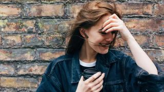 a woman wearing a pair of the jbl tune 225tws true wireless earbuds, she's laughing and stood against a brick wall