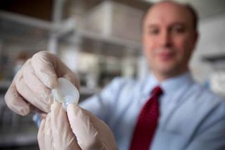 A scientist is shown holding up a 3D-printed ear to the camera. The ear is in focus but everything else is blurred.