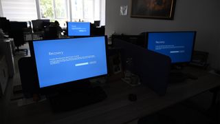 Microsoft outage image showing blue screen of death on multiple devices in an open plan office space.