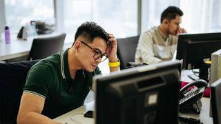 Stressed IT admin working at a computer station in an open plan office space.